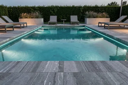 Piscine extérieure moderne avec éclairages LED sous-marins bleus au crépuscule, reflets sur l'eau calme et dallage en travertin gris